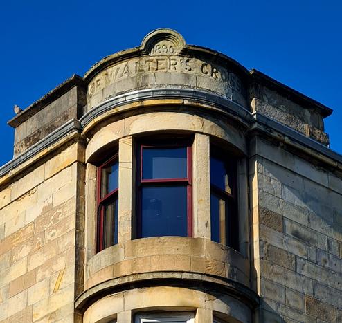 The top of a column of semi-circular corner windows with the words Sir Walter's Cross and the date 1890 carved into its pediment.