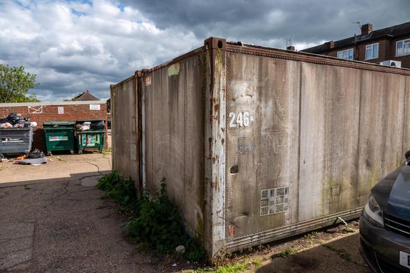 An ISO Container has '246A' stuck on the side of it. Behind it are overflowing rubbish containers, and above it are windows and chimneys of houses.
