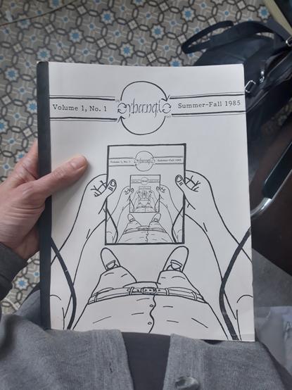A hand holding a copy of the "Volume 1, No. 1, Summer-Fall 1985" issue of the "Cybernetic" Zine. The cover is a recursive mise en abyme of hands holding the cover of a zine, with a picture of hands holding the cover of a zine, etc.