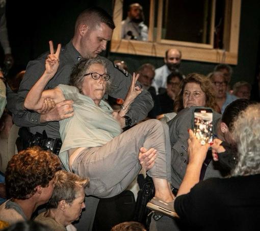 An elderly woman is being physically removed from a crowded room by a security guard. She is holding up both hands and flashing the victory sign.