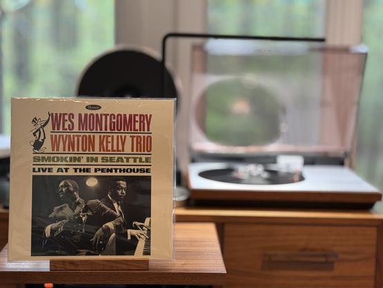 Wes Montgomery / Wynton Kelly Trio - Smokin' in Seattle: Live at the Penthouse LP over.

Off-white background, artists and title at the top in large font, with the Penthouse club mascot leaning up against the artist names.

A black and white shot of Wes (left) and Wynton (right) is below, taking up the bottom half.

The black LP plays on a vintage, restored Bang & Olufsen Beogram 4000 linear-tracking turntable to the right.
