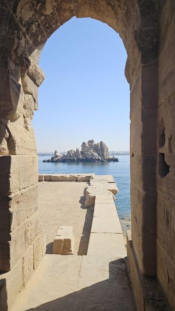 Photo looking through a narrow arch of a temple ruin. Behind the arch is a low foundation where once was a building. The sun shines across the flat stone, but doesn't reach me standing in the shadow of the arch. Behind the ruins lies the blue Nile water and not far away is a tiny island made entirely weird rock formations. Above the water is the blue sky which is not quite as blue as the Nile water. The scene looks calm and peaceful.