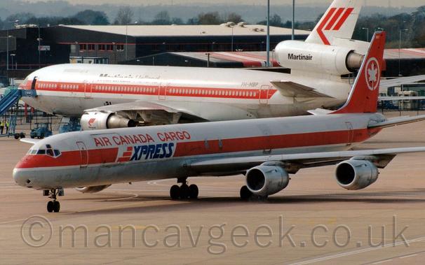 Side view of a long and thin, white and red, 4 jet-engined cargo plane taxiing from right to left.
The plane is mostly white, with a grey belly, and a thick red stripe running along the body.
There are red "Air Canada Cargo" titles on the upper forward fuselage, with blue "Express" titles just below.
The tail is red, with the white outline of a circle, with a white maple leaf in the middle.
Grey concrete apron fills the foreground.
A white, 3 engined jet airliner with a red stripe running along the body and black "MartinAir" titles on the centre engine is in the background, with black buildings beyond that, under grey skies.
