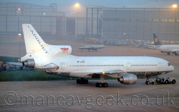 Side view of a white, 3 engined jet airliner facing the right but being pushed back to the left by a low-slung blue and grey tug attached to the nosewheel.
The plane has a grey belly, with blue "CaribJet" titles running diagonally down the leading edge of the tail.
The registration V2-LEO is on the upper rear fuselage in black, and there are red "TriStar 500" on the centre engine intake.
2 men in hi-viz jackets are standing between the plane's nosewheel and the tug.
Grey concrete apron fills the foreground, as well as a large chunk of the background, partly separated by a grassed are on the left of the frame.
A pair of large, grey hangars dominate the background, with a pair of other planes parked in front - a grey and almost black twin propellor-engined airliner on the left, and a white twin engined jet airliner with a dark blue and yellow tail on the right.
It is obviously dusk, the whole scene being illuminated by bright lights, giving the whole image a slight yellow cast, with grey-blue sky visible between the hangars.
