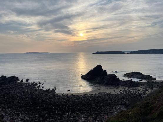 A calm sea with the low sun reflected through hazy clouds in the still water. There are dark rocks on the shore in the foreground and islands in the distance.