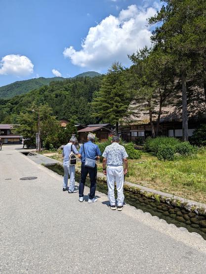 Three older men looking at the fish that swim in an human-made stream of water that passes a small hamlet. They are dressed in white and blue colors. In front of them, past the stream, there are trees, traditional Japanese houses and forested mountains.