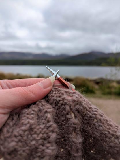 1) A photo of a hand holding up a purple knitting WIP in front of a blurry view of a loch and hills. The knitting is on the wrong side but you can see the backs of cables and lace work.