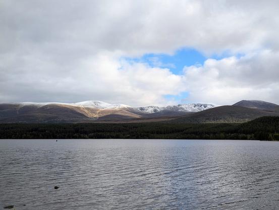 2) The loch and hills that were blurred in the background of the first photo. The tops of the hills have snow on, above brown/green hillsides. The loch is dark with some ripples. The sky is cloudy with some bright blue peeking through.