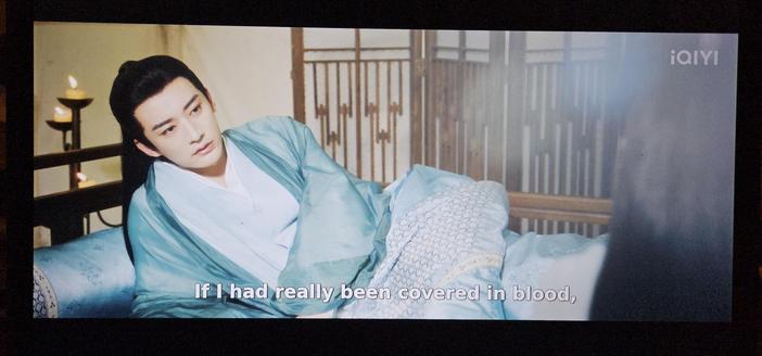Yuanzhong, in turquoise clothes, resting in a bed with turquoise sheets and pillow. The pillow fabric is woven in a very common chrysanthemum pattern, and the bed sheet have a symetrical ovals surrounded by a corresponding net pattern woven into it.