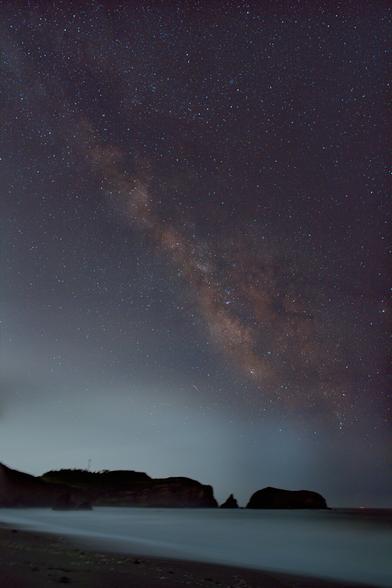 The Galactic Center of the Milky Way points up and away from the horizon. Thousands of stars are visible, and the Milk Way has a reddish hue. In the foreground is a beach at night with cliffs in the distance. The surf is silky white. There is a glow on the horizon from city lights.