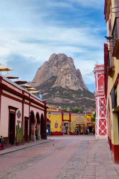 A empty street, with paving stones, surrounded by one-story colourful buildings, with a big rock, boulder, mountain in the background. during a cloudy day