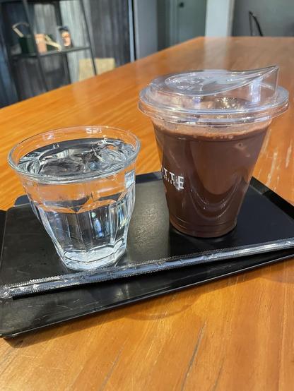 A plastic cup of chocolate drink and a glass of water sit on a black tray with a straw, atop a wooden table.