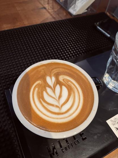 A latte with latte art is on a black tray. The tray has the words "Miitte Coffee" in white lettering. A glass of water is next to the tray.