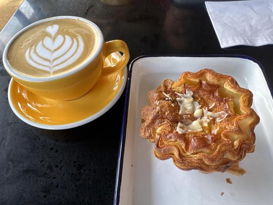 A black metal outdoor table supports a handsome latte in a buttery orange/gold cup and saucer, sporting the traditional stacked hearts of a well-made coffee drink.  It was quite good, if not exactly hot enough for Teflon-brain.

On a white squarish plate to the right of the coffee is a small "quiche" which was a tiny amount of an egg mixed with cheese baked in the center of a pastry cup made out of perhaps puff pastry?  It is all a uniform orangey-brown.

It was an excellent recovery, from a 35 minute drive that might have ended in hunger.