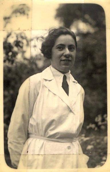 A woman in a white coat and tie is standing. Some plants are behind her. You can see her fro, the waist up.