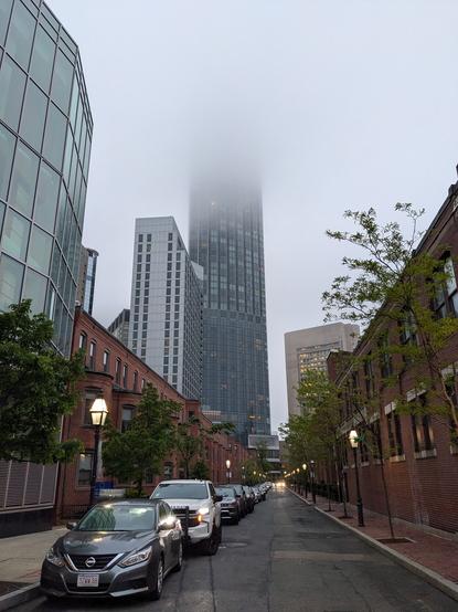 The residential tower next to the Prudential center in Boston, as seen from the intersection of Mass Ave and St Germain St.  It is foggy and the top of the tower can't be seen in the clouds.  The road is wet.