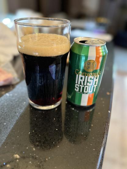 A pint glass of dark beer next to a shiny green can with a white and orange vertical stripe. The gold logo is a circle in the middle