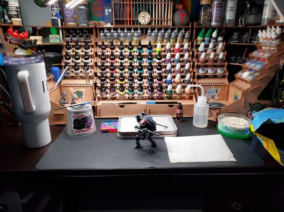 Photo of a desk covered in miniature painting supplies, a partially painted Imperial Knight, some drinks, and some snacks.