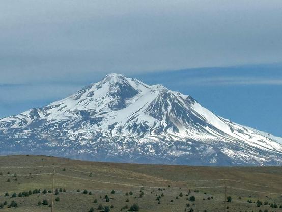 A large snow covered volcanic mountain covers the entire horizon with a lower rolling green-gray hillside dotted with bushes that might be tumbleweeds. The sky is hazy.