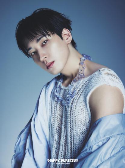Wonwoo poses in a fashion-forward outfit featuring a knitted top and a light blue jacket. He wears a unique necklace made of transparent pieces and has short, styled hair. The background is a soft blue gradient, and the image is captioned "Happy Burstday”