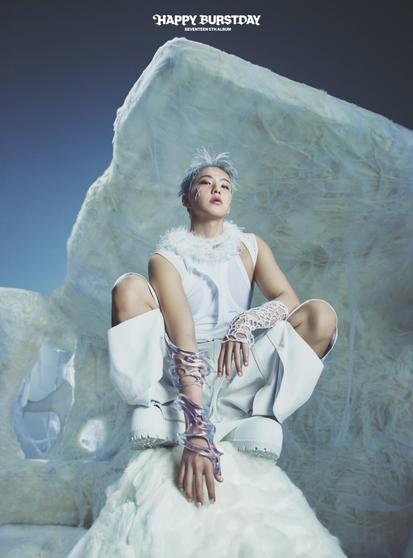 Hoshi with short, silver hair sits on a fluffy white surface, wearing an all-white outfit featuring a sleeveless top, loose pants, and unique, transparent accessories on his arms. The background has a stylized icy landscape.