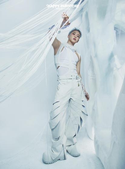 Hoshi poses in a futuristic white outfit with unique designs, including a sleeveless top and layered baggy pants. The setting features light, ethereal colors and textures, with strands of white material in the background. The image conveys a stylish and artistic vibe.