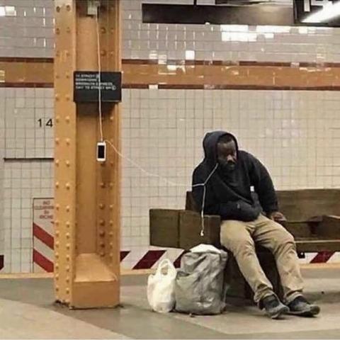 A person charges their phone from a socket on a pole in an underground station. The headphone lead stretches from their dangling phone to where they sit on the nearby bench.