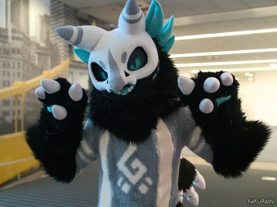 A gray fursuiter with black hands and white claws, stubby bony wings, and a white skull-like pattern on the head, as if wearing it as a helmet. Cyan feathers poke out behind the head. The eyes and tongue are cyan as well. Stripes run across the upper arms and the chest. There is a white emblem on the abdomen. In the left of the background is a grayscale photo of the Pittsburgh downtown skyline, with only a cable-stayed bridge highlighted in yellow. A striped carpet on the floor mirrors bars of ceiling lights, both receding into the distance.