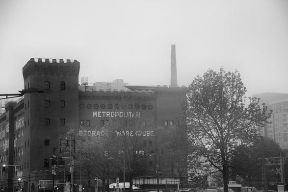 A black and white photo of a historic warehouse building with a wall displaying the words "METROPOLITAN STORAGE WAREHOUSE" is shown, partially obscured by mist. A tall smokestack rises in the background, and there are trees and traffic signals in the foreground