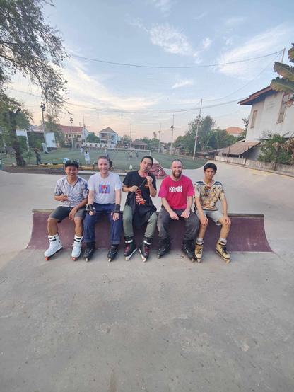 Five men – two white, three Asian – wearing inline skates sit on an obstacle at a skate park. In the background a football ground and South East Asian architecture is visible.
