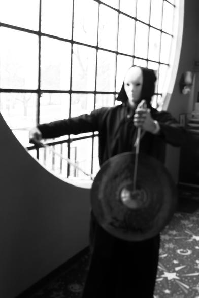 A figure in a black hooded robe and a white mask holds a gong and a mallet in a room with large circular windows.