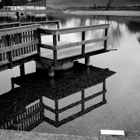 The black and white image shows a wooden dock extending over a calm body of water, with its railings and platform clearly reflected in the still surface below.
