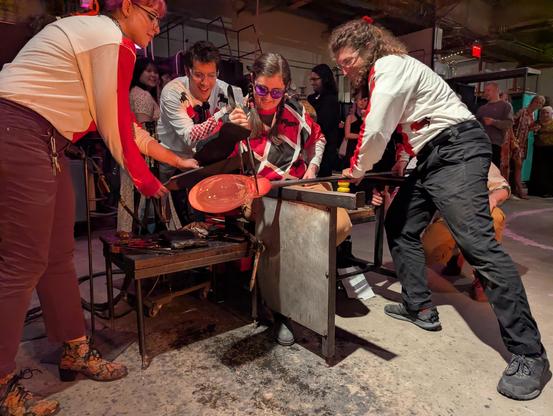 Ali blowing glass- putting the jackline I to a large bubble - the horse trophy. Other glassblower performers dresses in horse jockey shirts assist, turning the large blowpipe and shielding Ali's arm with blackened wood paddles from the radiant heat of the bubble