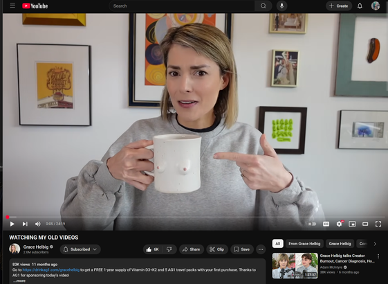 Screenshot from her video "WATCHING MY OLD VIDEOS" showing her pointing to a ceramic mug with breasts on it while arching her brows in bemusement