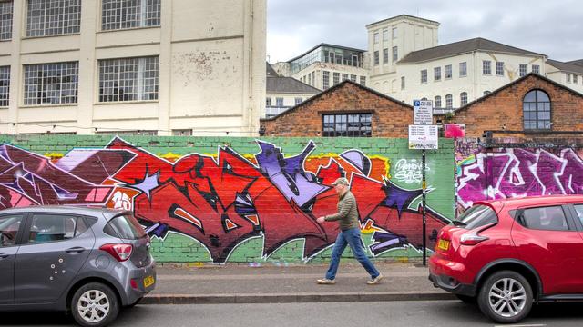 A man walking past a wall painted with street art / graffiti. It's shot from the other side of the street and the man is visible between two parked cars. He's walking left to right with his arms swinging. The wall behind him is painted green with red green blue yellow and purple abstract shapes. Behind the wall, run down industrial buildings are visible.