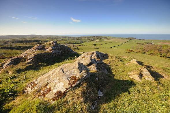 Photo of a rocky outcrop in the foreground, with a view over green fields to the sea, under a blue sky.