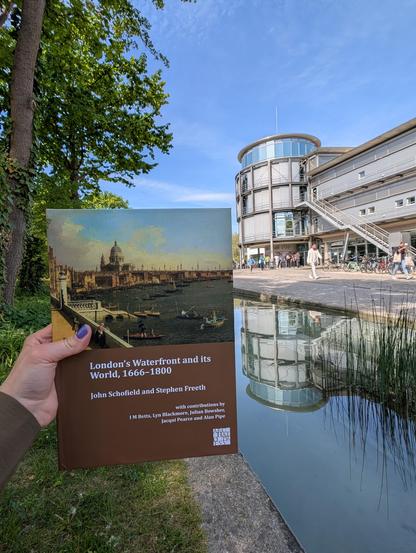 Holding John Schofield and Stephen Freeth's book "London's Waterfront and its World, 1666-1800" in front of the water course in front of SUB Göttingen. Behind the book, the round cafeteria tower of the library is visible