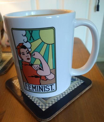 Mug of tea on a bedside cabinet, with a picture of Rosie the Riveter dressed in red, wearing a red bandana, her right fist raised, above her to the left is a white cloud, to the right is a yellow sun, with green and yellow rays. Underneath is the word Feminist.