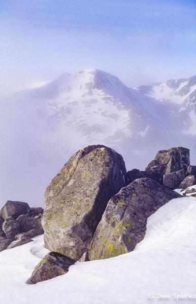 In the foreground, grey boulders surrounded by snow on a mountain ridge. In the background, seen through a light mist, two more snowy peaks.