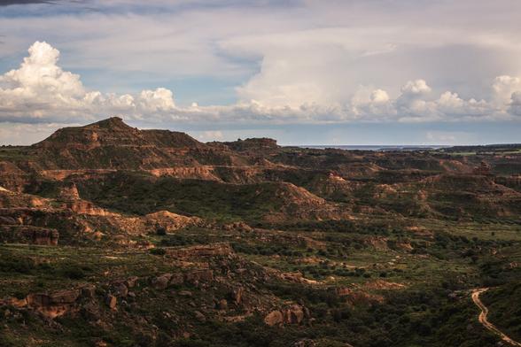 Photograph showing a semi-desert landscape, with limestone hills and some green vegetation. Above this landscape, a blue sky with plenty of clouds.