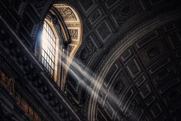 Sunlight streams through a large arched window into a dimly lit interior with ornate, patterned ceilings and walls inside Saint Peter's Basilica in the Vatican.
