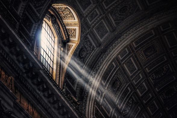 Sunlight streams through a large arched window into a dimly lit interior with ornate, patterned ceilings and walls inside Saint Peter's Basilica in the Vatican.