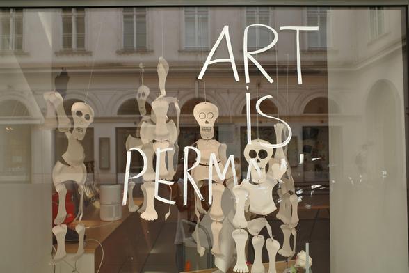 The image shows a storefront window display featuring white skeleton-like paper figures hanging by strings, with the handwritten phrase “ART IS PERMA” written in large white letters across the glass. The reflection of a building with arched windows is visible in the background.