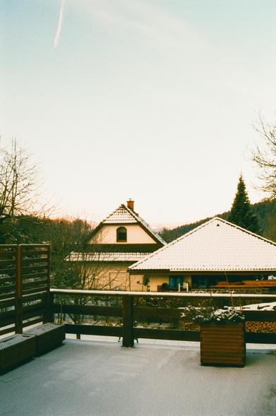The image shows a snowy winter scene with a view from a deck or balcony. In the foreground, there are planters and a railing covered with snow. In the background, there are two houses with snow-covered roofs. The sky is clear with a contrail, indicating an aircraft passed by recently. Bare trees are also visible, suggesting a winter setting.