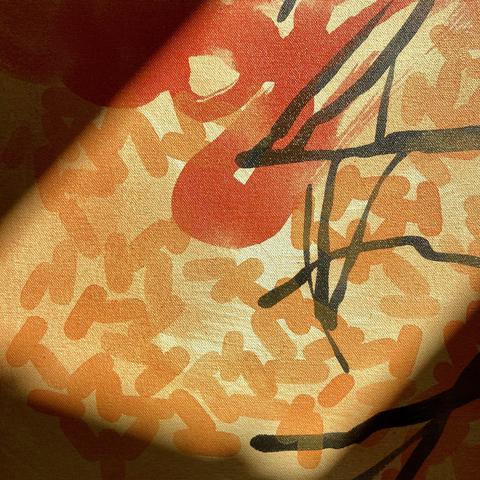 A close-up detail of an acrylic painting on canvas that is illuminated by a sunbeam. The detail is of inky burnt tree branches on a pale orange background, gold-ish swarms of lozenge shapes, and a pale red semi-transparent blob that is in the center.
