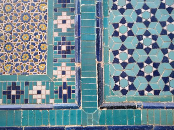 Two sets of hexagonal tiles separated by a plain border. 
The left set of tiles: interlaced white lines make hexagons and hexagrams containing yellow flowers. Red, blue and yellow polygons fill in the other areas. The hexagon tiles have a middle border of white, cyan and blue square and rectangle tiles making cross shapes. 
The right set of tiles: the centre of each tile is a cyan hexagon surrounded by white equilateral triangles and blue kites.