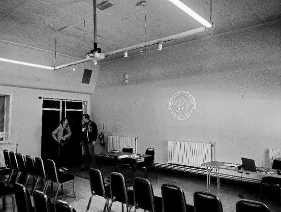 A white/black photo with two people, empty room, and logo of the conference presented on the wall