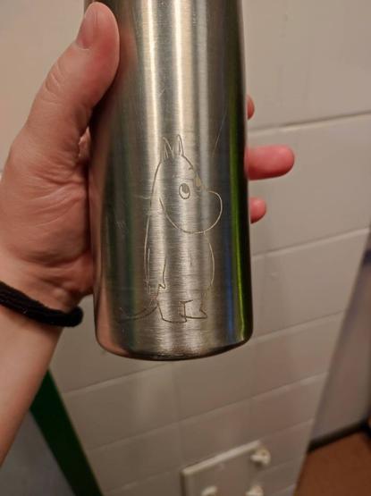 Hand hält eine Edelstahlflasche mit einem eingravierten Moomin-Logo, Nilpferdähnliche Trollwesen