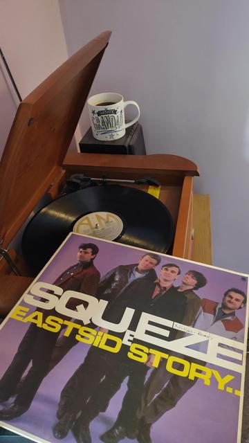 Record player with a 12" black vinyl record.
In the foreground is an album cover showing the five band members standing in front of a violet background.
Behind the player is a mug of coffee on a speaker.