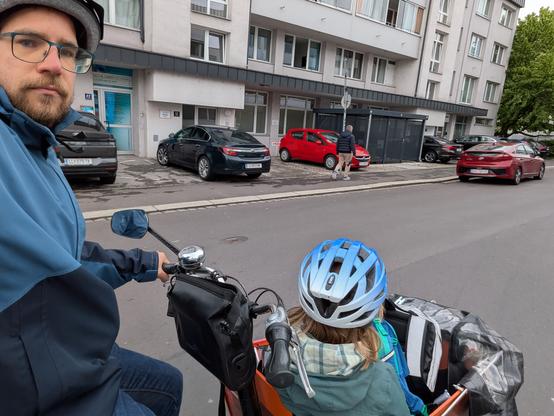 Das Bild zeigt einen Mann, der auf einem Lastenrad mit Kind in der Transportkiste fährt. Der Mann trägt eine blaue Jacke und eine Brille und hat einen Bart. Das Kind im Kinderwagen trägt einen blauen Radhelm und eine grüne Jacke. Im Hintergrund ist ein mehrstöckiges Wohngebäude mit Parkplätzen zu sehen, auf denen mehrere Autos geparkt sind, darunter ein rotes Auto.Die Szene wirkt ruhig und alltäglich, mit einem typischen Stadtbild im Hintergrund.